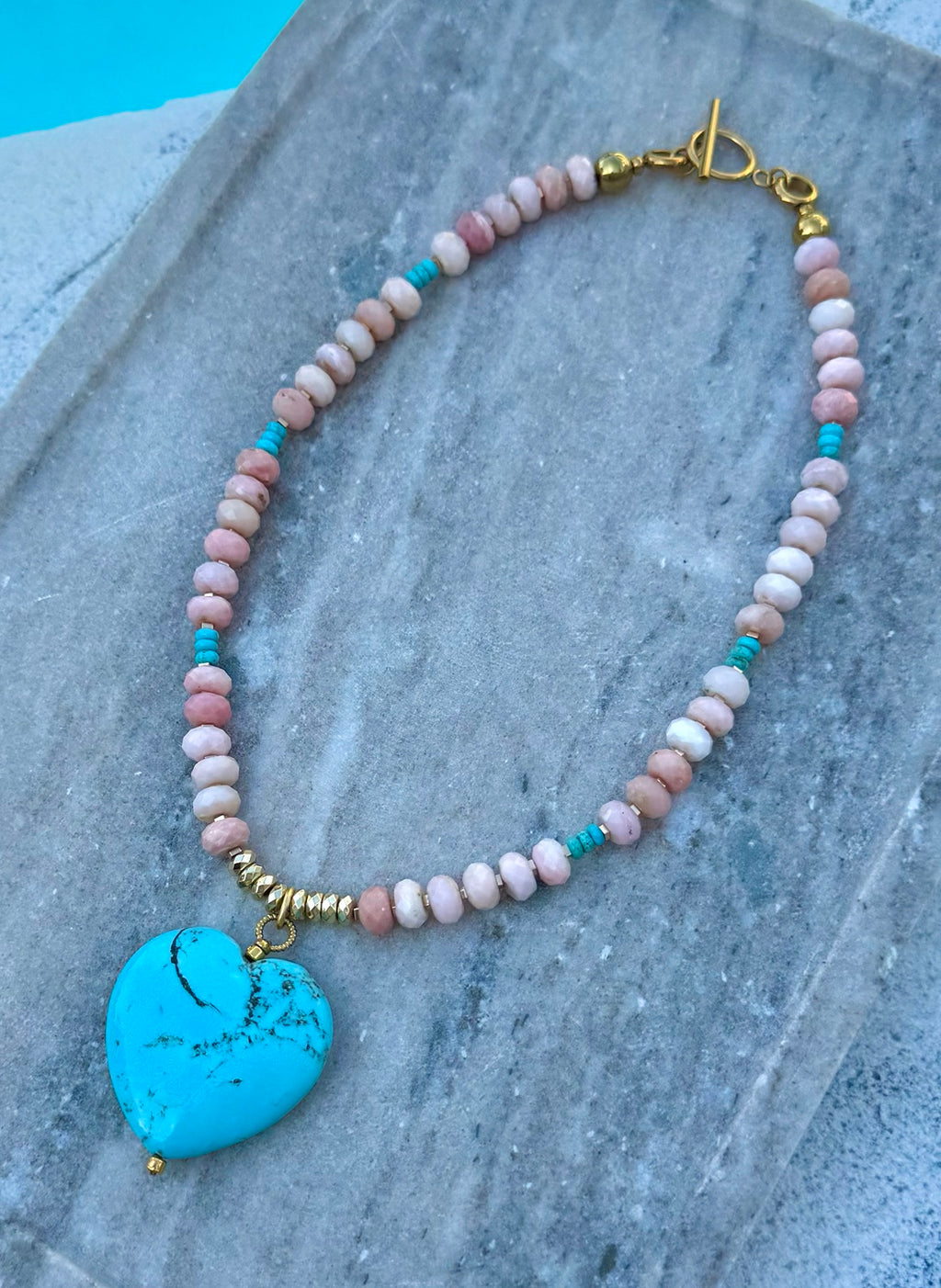 Pink opal necklace with turquoise heart pendant laid flat by the pool