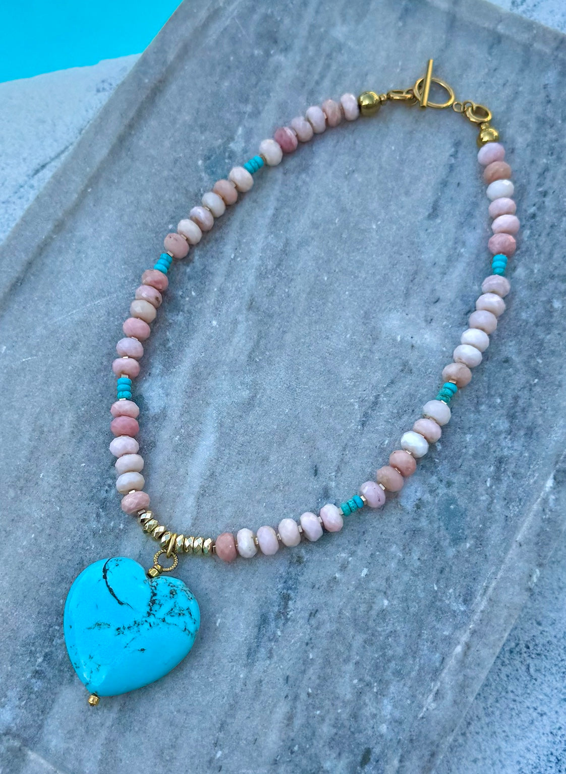 Pink opal necklace with turquoise heart pendant laid flat by the pool