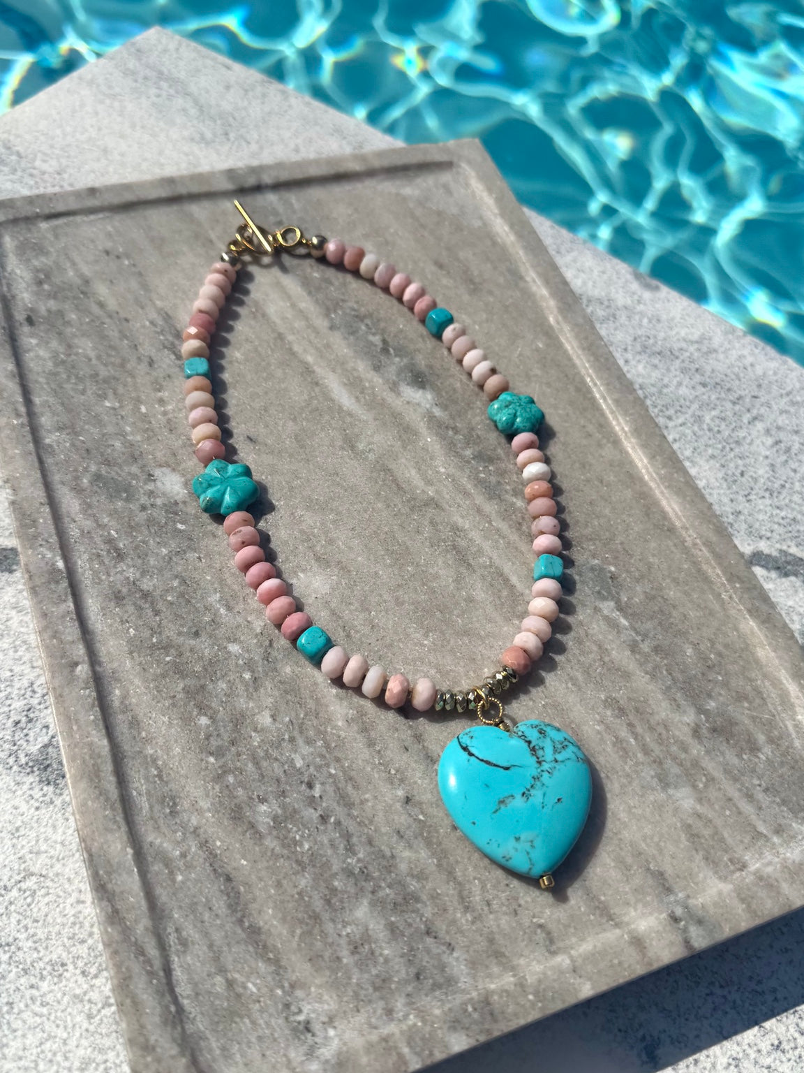 Angled poolside view of The Colt pink opal and turquoise flower heart necklace